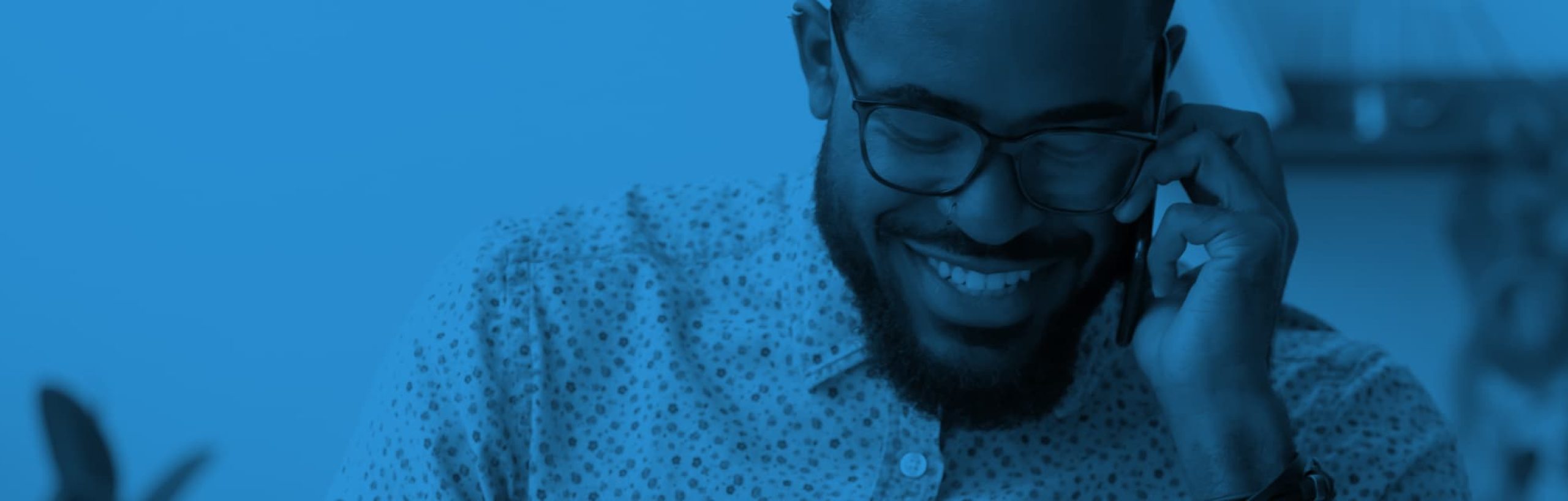 Part of a photo: a man wearing a dotty shirt is sat slightly off centre, talking on the phone, he has a broad smile and is looking down; there . The image has a blue filter.